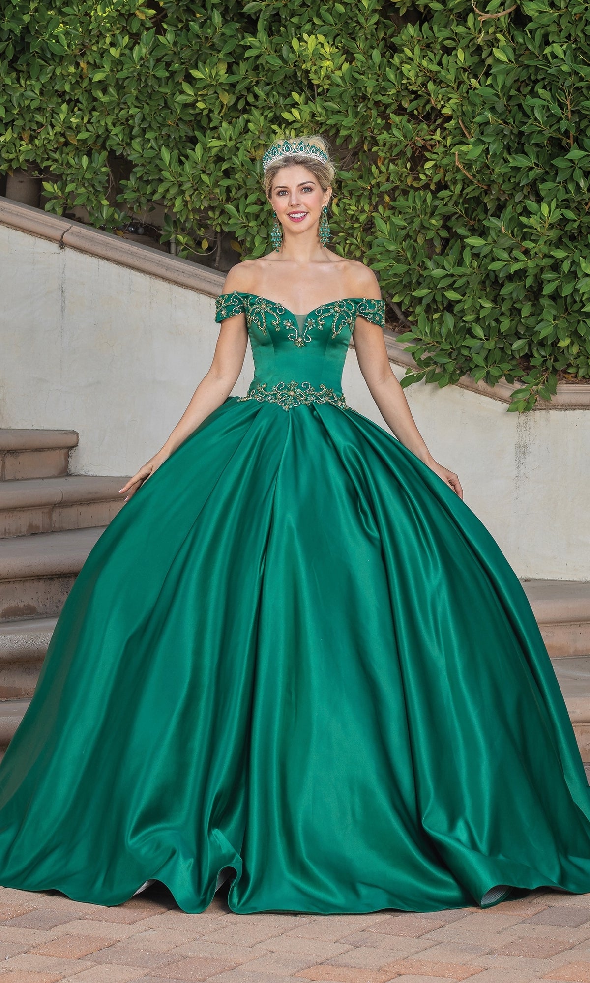 A woman stands outdoors near steps wearing Quinceanera Dress 1466 by Dancing Queen—an off-the-shoulder, emerald green A-line ball gown with a full skirt and a matching tiara.